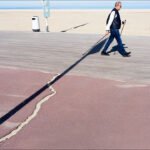 Man walking with cane on sunny seaside boardwalk with ocean view.