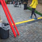 Person walking on cobblestone market pathway with wooden shops and bright yellow safety mat.