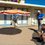 Sunny motel courtyard with orange beach umbrella, blue doors, flowers, and relaxing people.