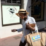 Stylish person in straw hat walking past Coco Chanel fashion display on a sunny city street.