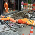 City construction workers in orange safety gear working in a street trench near a fashion boutique.