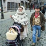 Elderly couple walking in city with Chihuahua in stroller on cobblestone street.