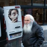 Elderly man with white beard in black leather jacket standing on wet urban street.