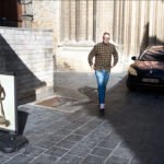 Man walking down sunlit cobblestone alley with statue and historic urban architecture.