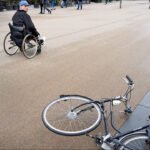 Man in wheelchair looks at fallen bicycle on city street.