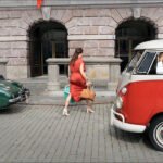 Woman in red dress walking past vintage cars on cobblestone street.