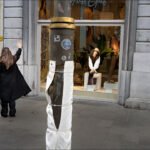 Woman in black coat waving near fashion store window with mannequins and pink handbag.