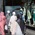 Women in hijabs walking past a city storefront displaying modern swimwear mannequins.