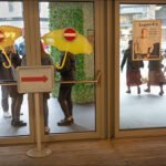 Colorful city scene with yellow umbrellas, glass doors, and people outside a modern storefront.